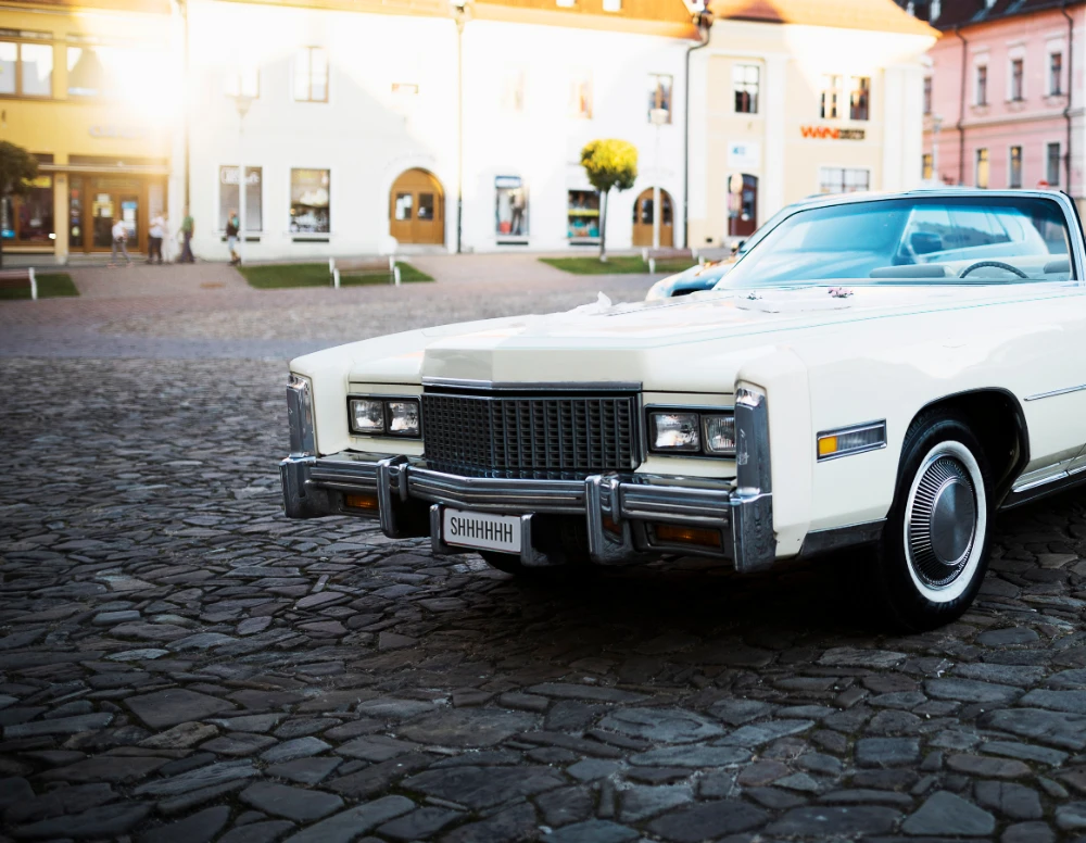 A vintage white Cadillac with a custom "SHHHHHH" license plate parked on a cobblestone street.