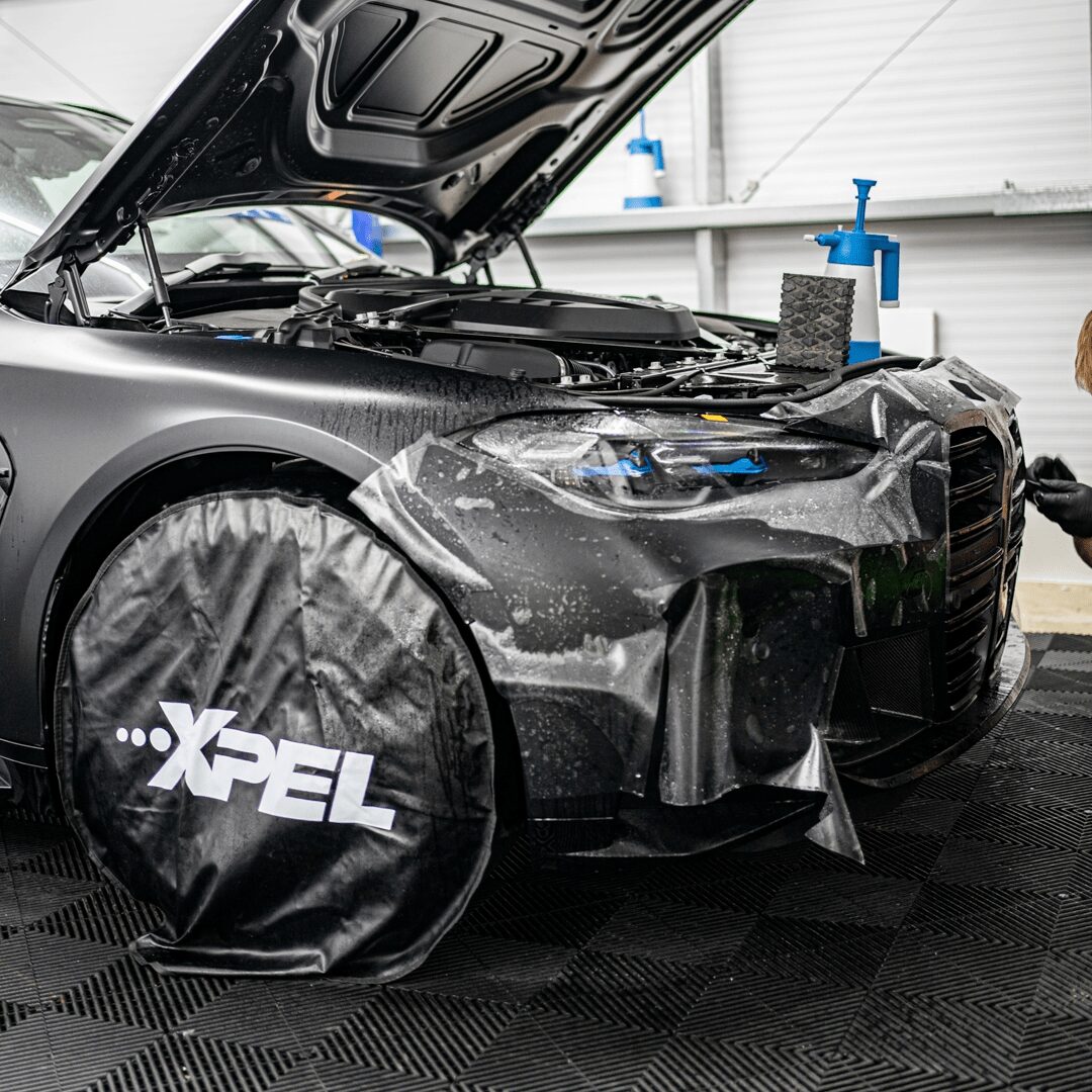 A black car undergoing a paint protection film (PPF) installation with "XPEL" branded wheel covers.