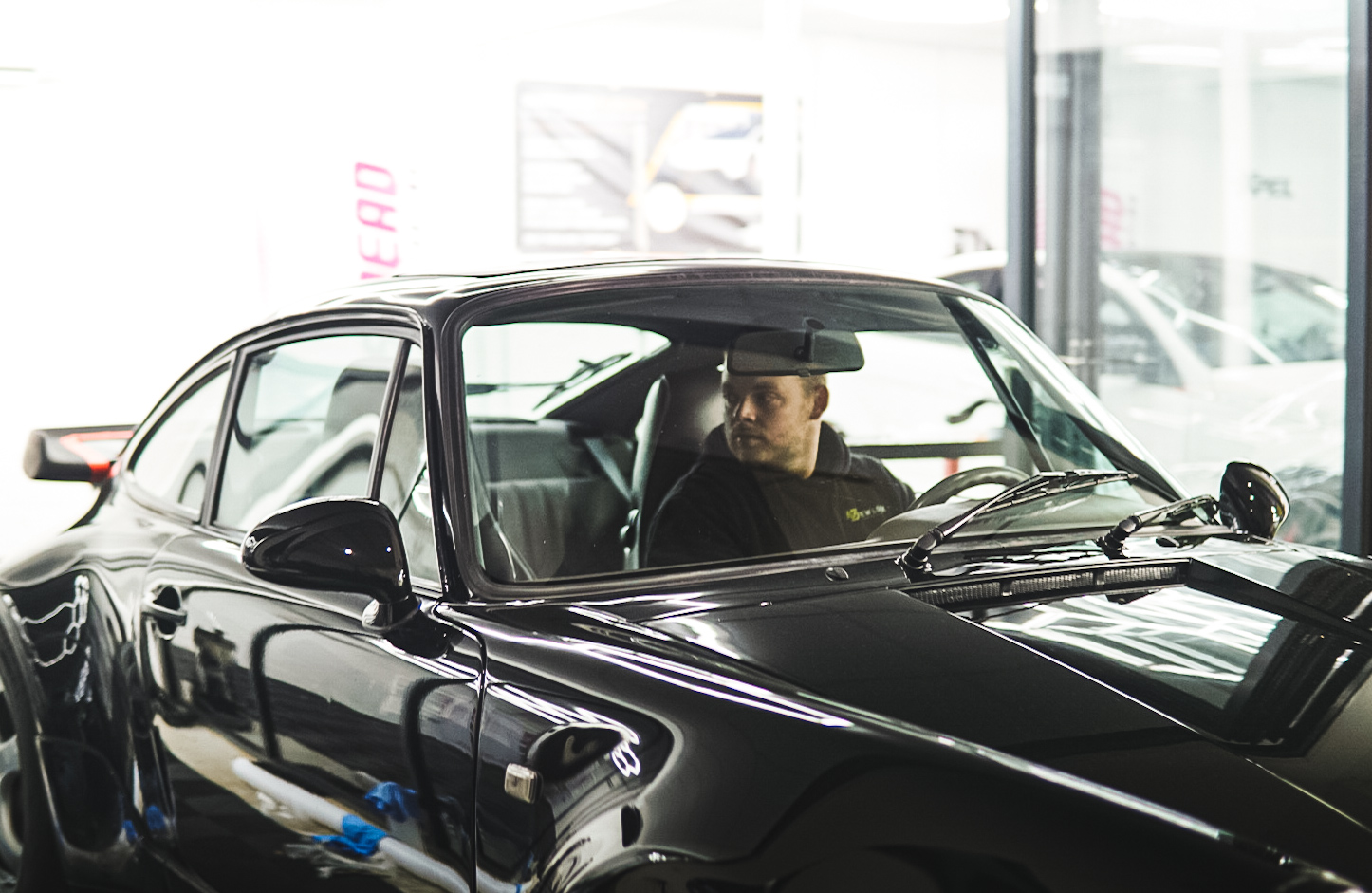 A detailer sitting inside a black Porsche with a glossy finish inside a workshop.