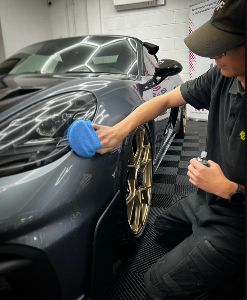 A detailer applying ceramic coating to a grey Porsche with gold wheels using a microfiber applicator.