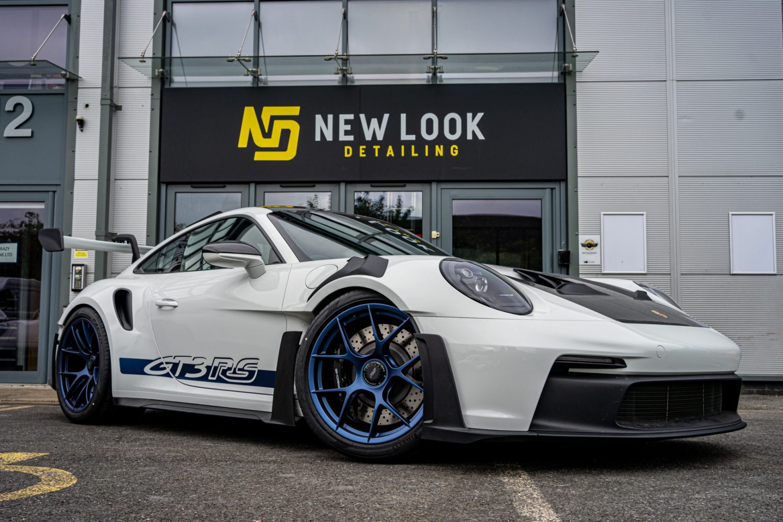 A white Porsche GT3 RS with blue wheels parked outside "New Look Detailing" studio.